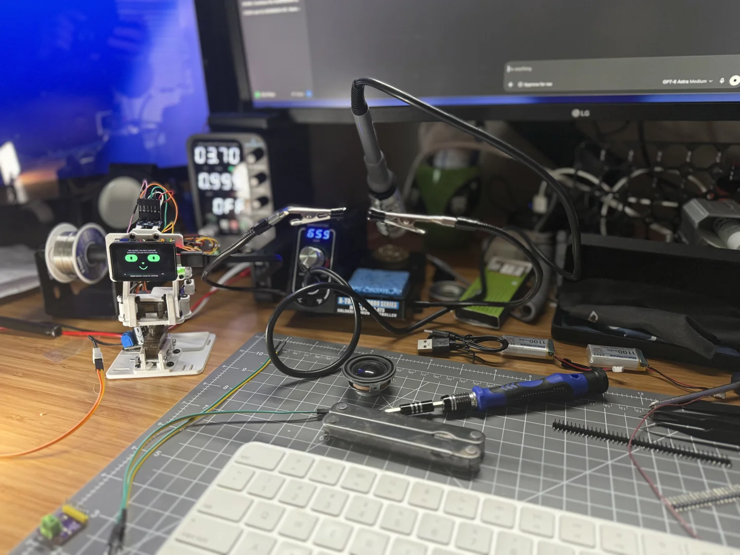 Photograph of the assembled grexBot prototype on a workbench, its green eyes lit, beside a soldering station, a loose speaker, and hand tools.