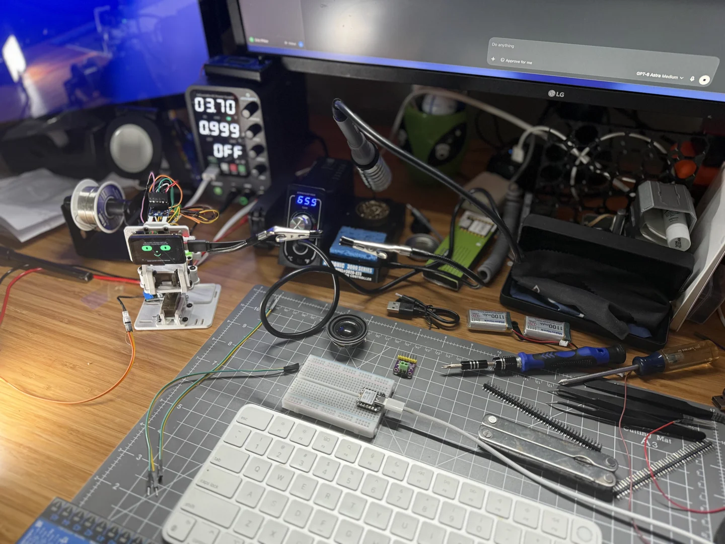 Wider photograph of the workbench, with grexBot, a cutting mat, a breadboard, and loose electronic parts